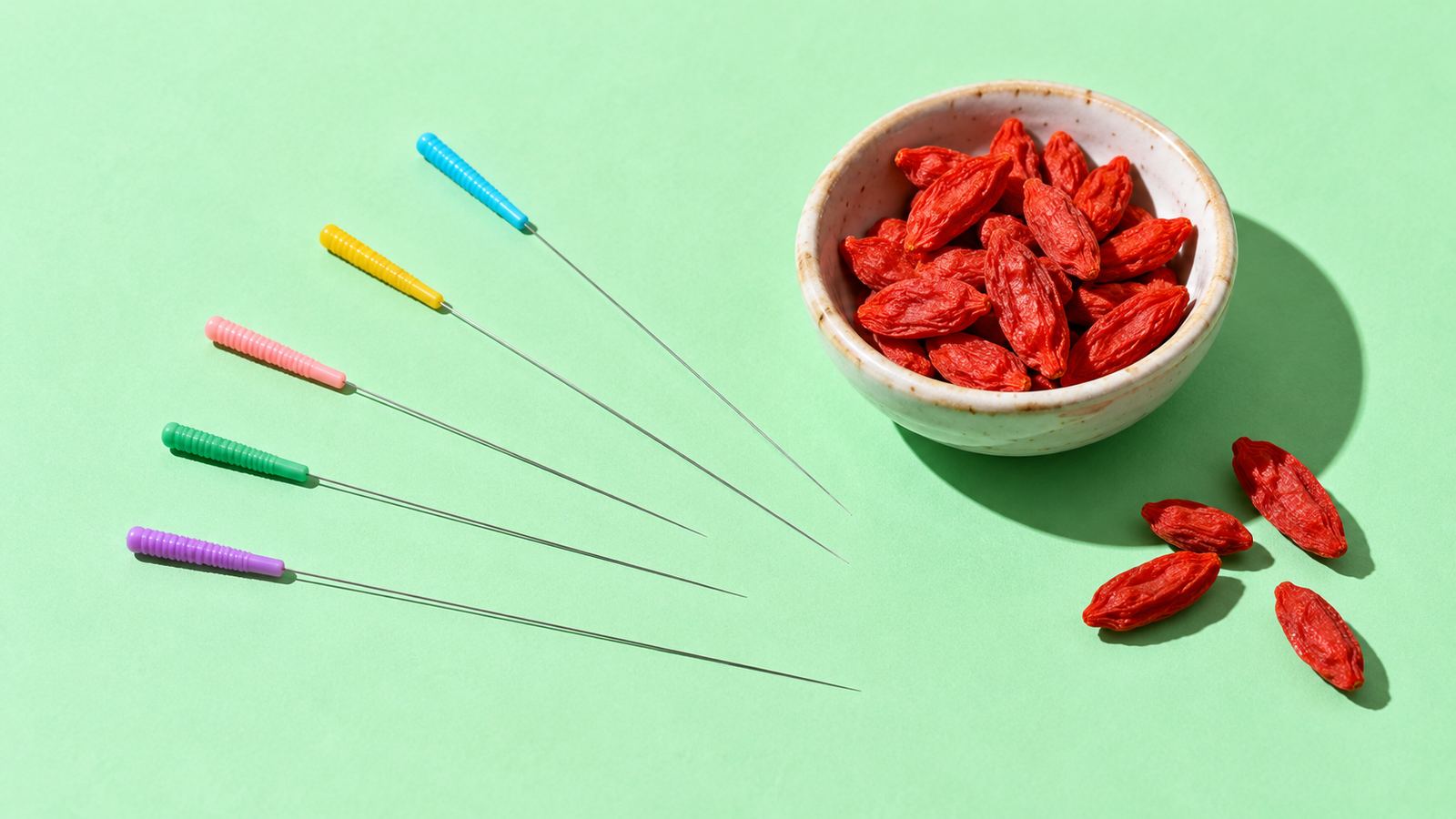 Five fine acupuncture needles with coloured handles beside a bowl of goji berries